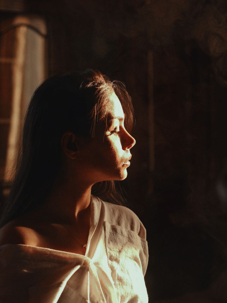Profile portrait of a woman turned towards a window, lit by warm late-afternoon light.