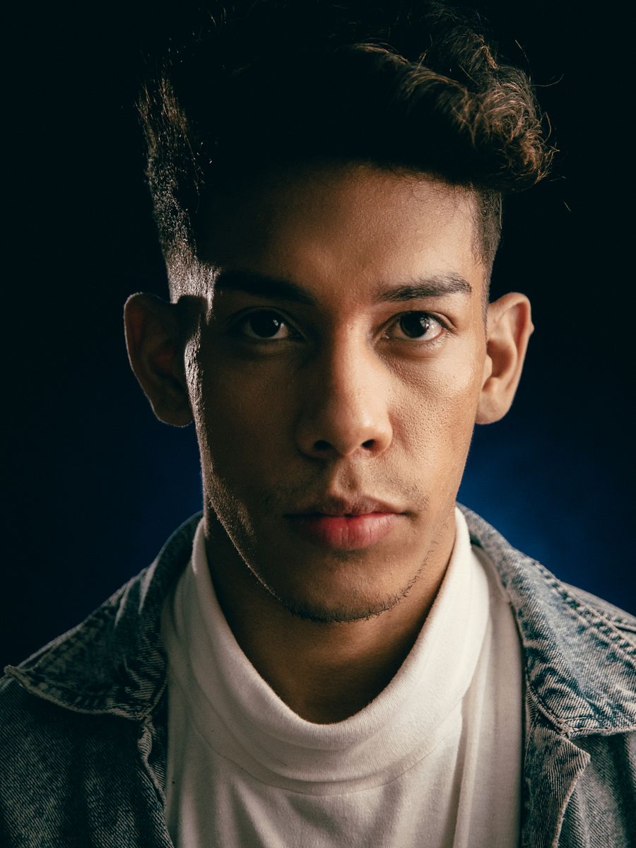 Portrait of a young man in a denim shirt, half his face in shadow against a dark backdrop.