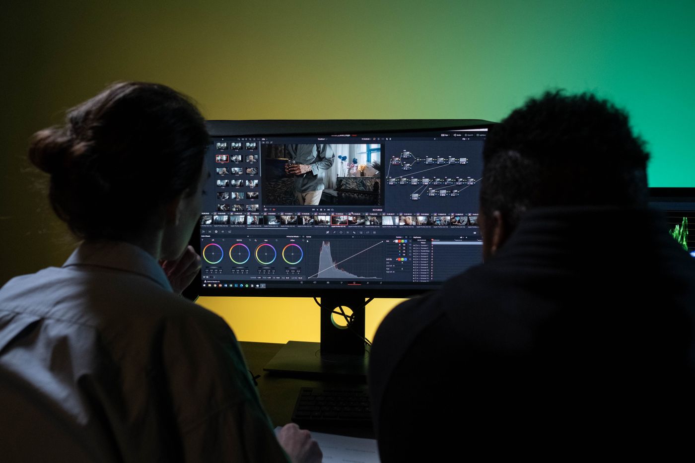 Two editors silhouetted in front of a monitor showing an edit timeline in a colour-lit suite.