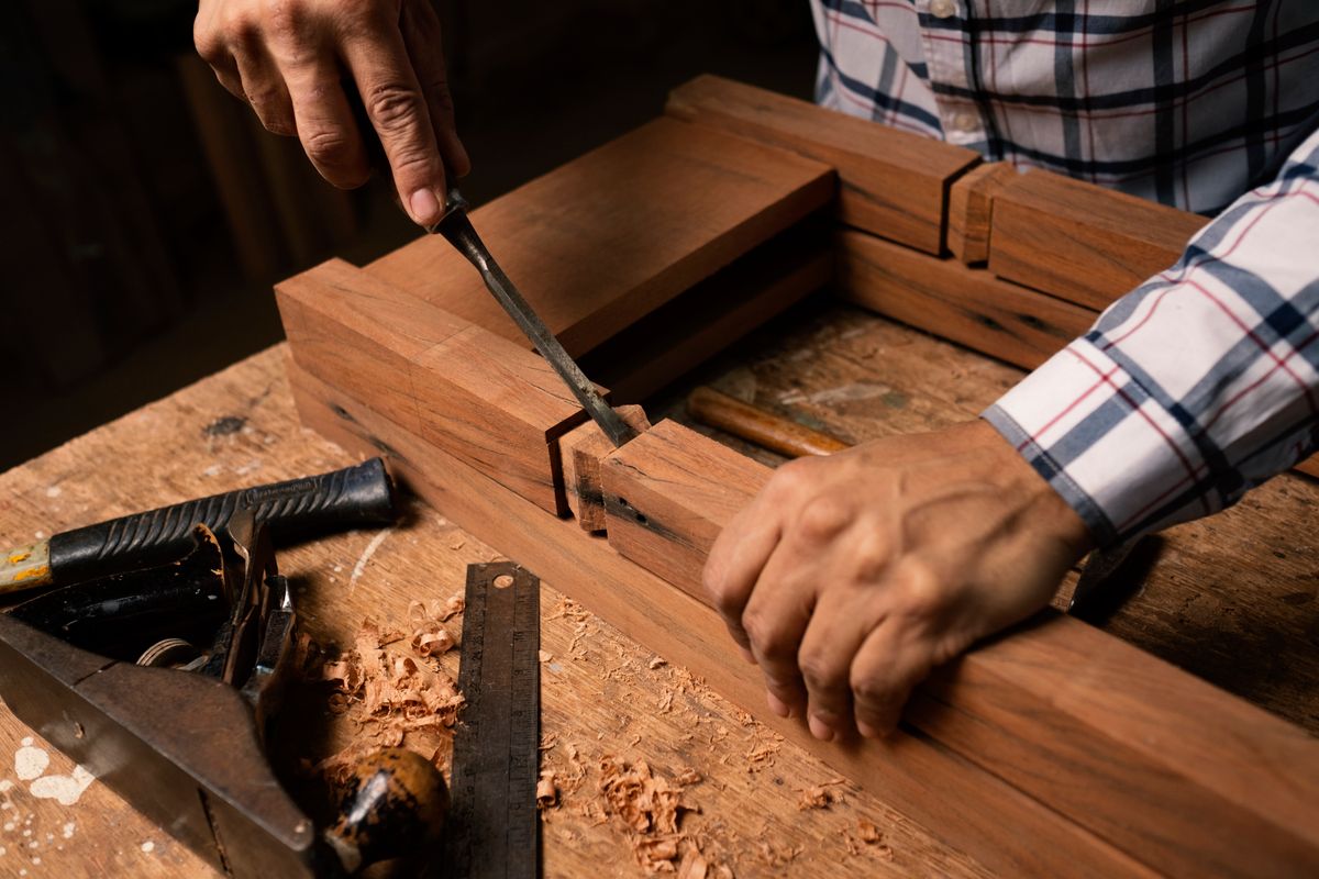 Close-up of hands cutting a mortise joint in a hardwood frame with a chisel, wood shavings scattered around.