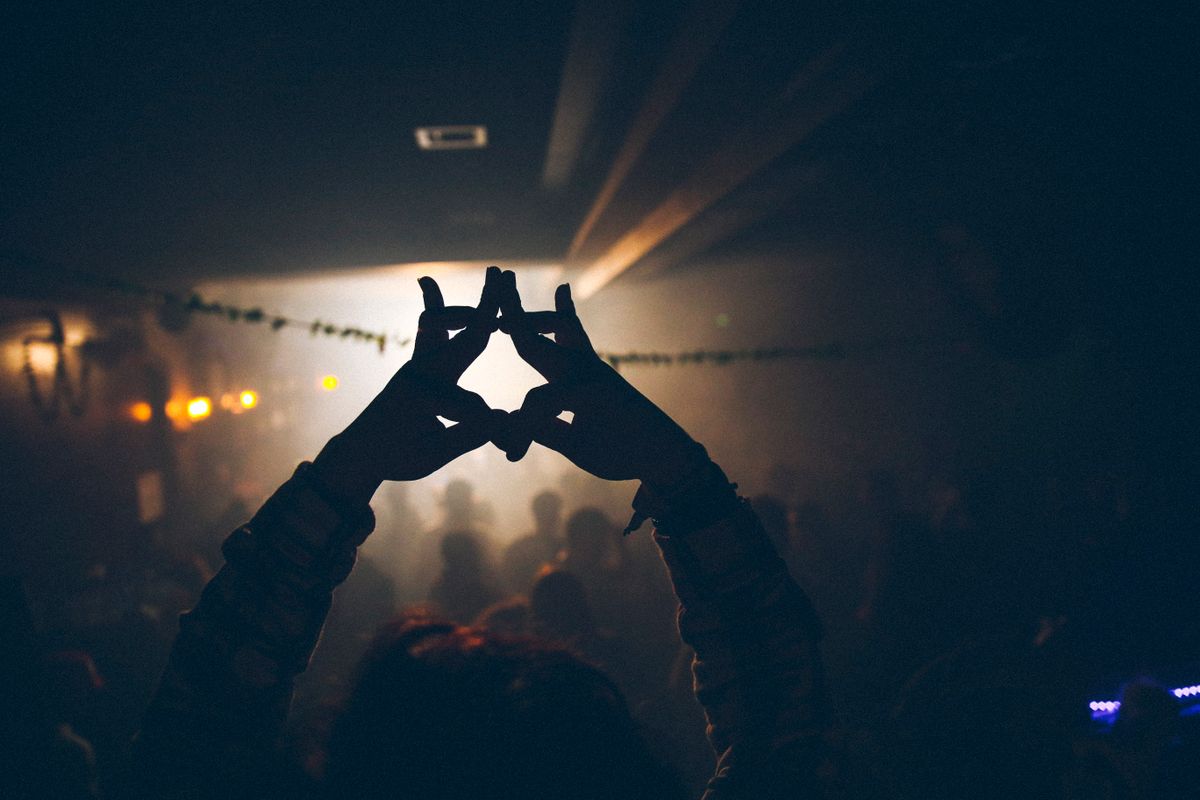 Silhouetted hands raised into a heart shape against warm stage lights at a night concert.