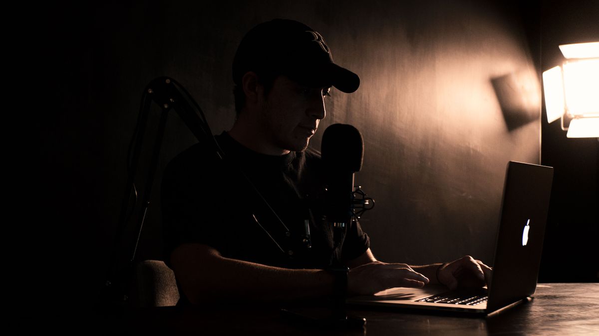 A person speaking into a condenser microphone beside a laptop in a warmly lit dark room.