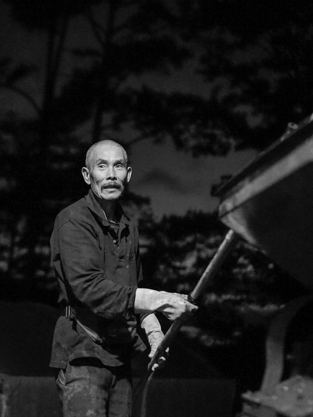 Black-and-white portrait of a coffee farmer standing beside a hulling machine at night.